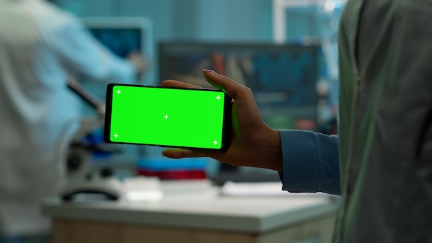 Microbiologist holding smartphone with green chroma key display in front of camera in modern equipped lab. Team of biotechnology scientists developing drugs using tablet with mock up screen.