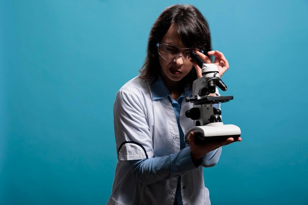 Microbiology expert using modern microscope to analyze virus at molecular level while on blue background. Chemistry specialist investigating chemical compounds inside substance sample with microscope.