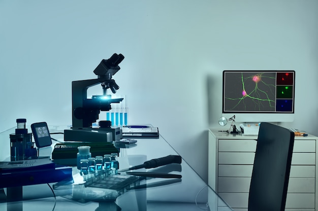 Microscoop, computermonitor met digitaal fluorescerend beeld van neuronale cellen en histologische fixatietools op glazen tafel.