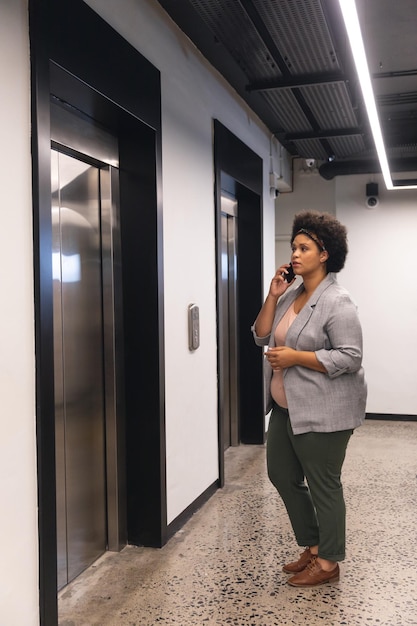 Photo mid adult biracial businesswoman talking on smartphone while waiting for elevator in office building