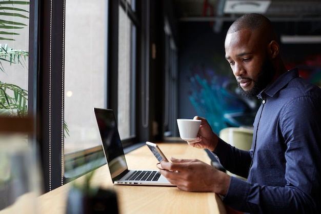 Mid adult black male creative sits by window having coffee using a laptop and smartphone side view