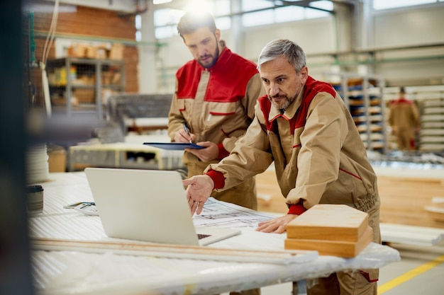 Mid adult carpenter using laptop while working with a colleague in workshop