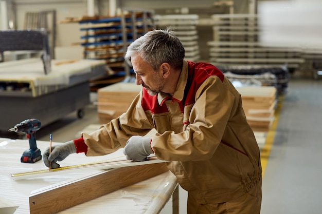 Mid adult carpenter using tape measure and marking wood plank in a workshop