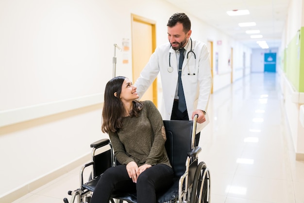 Mid adult doctor talking to discharged smiling young patient while pushing wheelchair at corridor in hospital