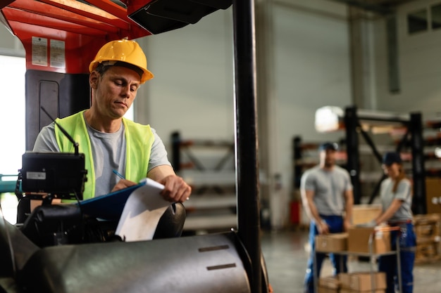 Mid adult forklift operator writing shipment reports while working in distribution warehouse