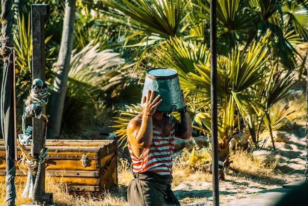 Photo mid adult man with bucket on head standing against palm trees