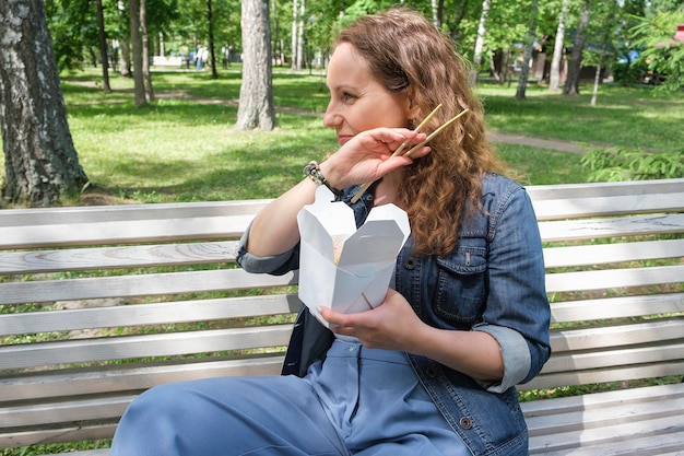 A mid adult mature woman on a walk on a summer day in the park has lunch with noodles wok