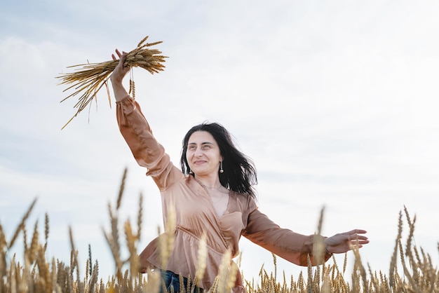 Женщина среднего возраста в бежевой рубашке стоит на пшеничном поле с восходом солнца на заднем плане