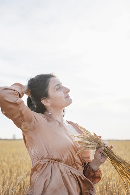 Mid adult woman in beige shirt standing on a wheat field with sunrise on the background back view