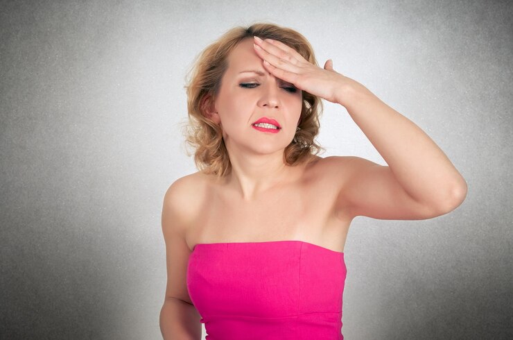 Mid adult woman in pink off shoulder dress suffering from headache against gray background
