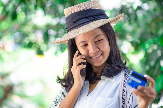 Photo mid adult woman with credit card talking over mobile phone while standing against trees