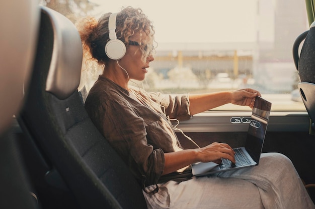 Mid age woman using computer inside transport bus vehicle during business or vacation travel Modern work digital lifestyle Female people with headphones enjoy transport Traveler lady with laptop