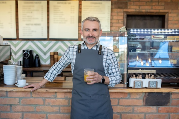 Mid aged man in apron standing near cafe counter with a glass\
of juice in hand