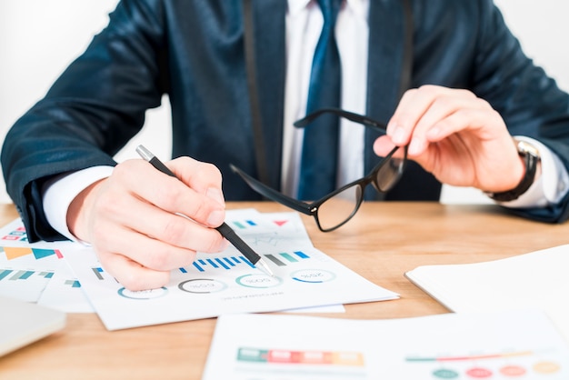 Photo mid section of a businessman holding black eyeglasses in hand analyzing the graph on wooden table