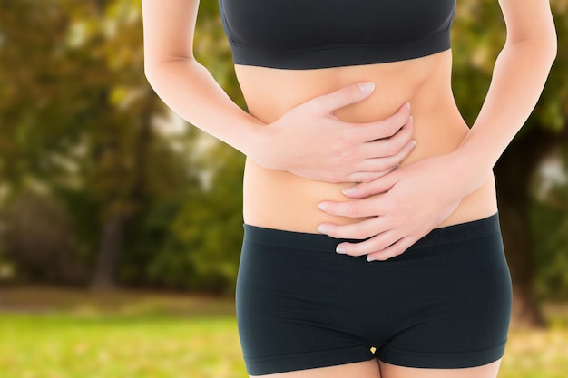 Mid section of a fit young woman with stomach pain against trees and meadow in the park