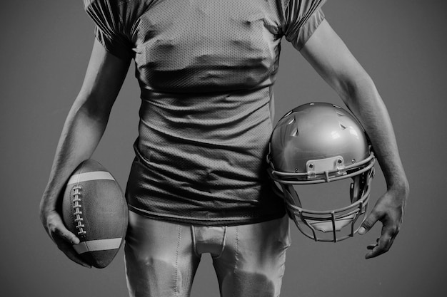 Mid section of sportsman holding American football and helmet against red background