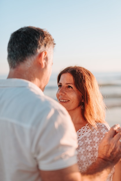 Mid volwassen paar op zoek naar elkaar ogen, witte jurken dragen op het strand tijdens een zonsondergang