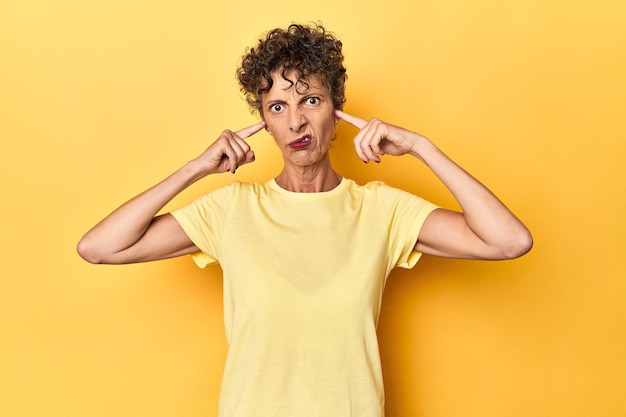 Midaged caucasian woman on vibrant yellow covering ears with fingers stressed and desperate