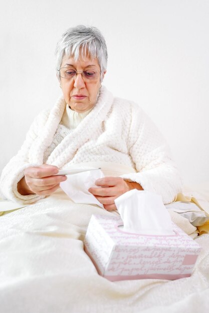 Foto midde volwassen vrouw die wegkijkt terwijl ze op bed zit tegen een witte achtergrond