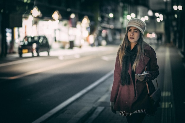 Midde volwassen vrouw die wegkijkt terwijl ze's nachts op een stadsstraat staat