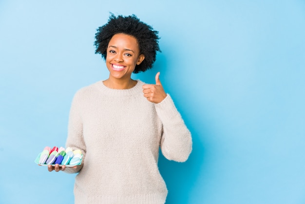 Middelbare leeftijd afro-amerikaanse vrouw eten bitterkoekjes geïsoleerd glimlachend en duim omhoog