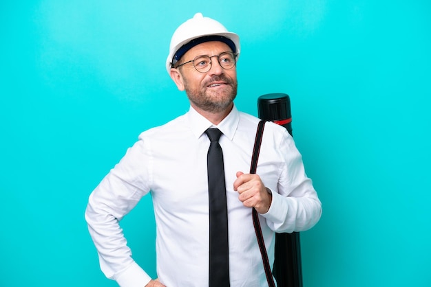 Foto middelbare leeftijd architect man met helm en houden blauwdrukken geïsoleerd op blauwe achtergrond poseren met armen op heup en glimlachen