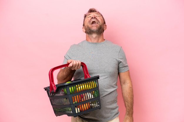 Middelbare leeftijd blanke man met een winkelmandje geïsoleerd op roze achtergrond lachen