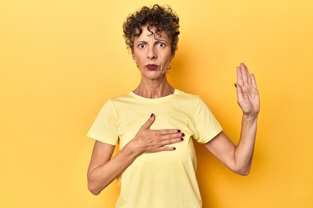 Middelbare leeftijd blanke vrouw op levendig geel die een eed aflegt en de hand op de borst legt