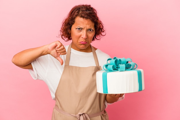 Middelbare leeftijd gebak Latijnse vrouw met een taart geïsoleerd op roze achtergrond met een afkeer gebaar, duim omlaag. Onenigheid begrip.