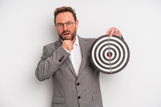 Middelbare leeftijd man met mond en ogen wijd open en hand op kin doel darts concept