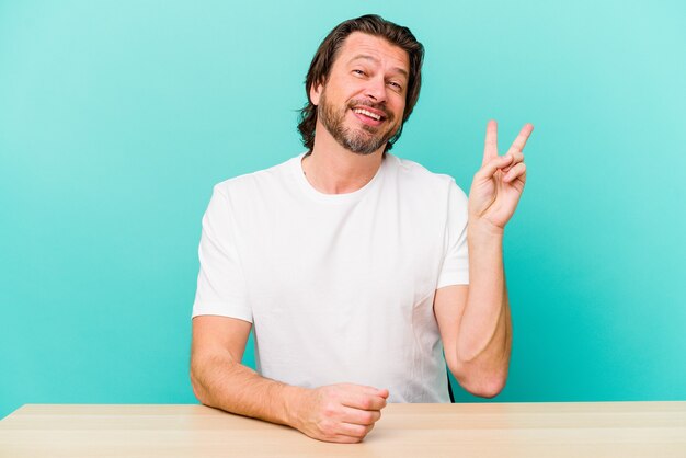 Middelbare leeftijd nederlandse man zit geïsoleerd op blauwe muur vrolijk en zorgeloos met een vredessymbool met vingers.