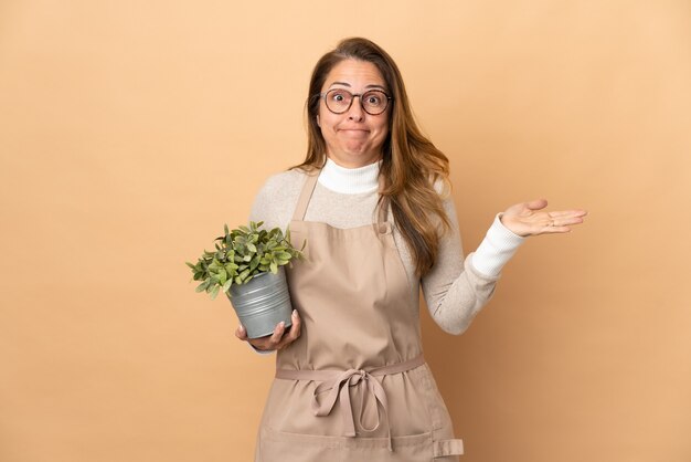 Middelbare leeftijd tuinman vrouw met een plant geïsoleerd op beige twijfels tijdens het verhogen van de handen