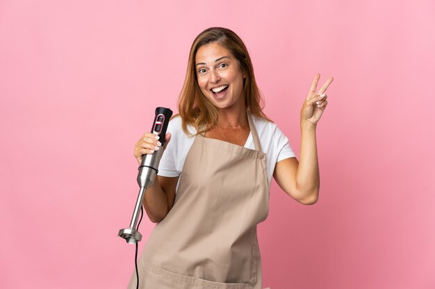 Middelbare leeftijd vrouw die handmixer gebruikt die op roze muur wordt geïsoleerd die en overwinningsteken glimlacht toont