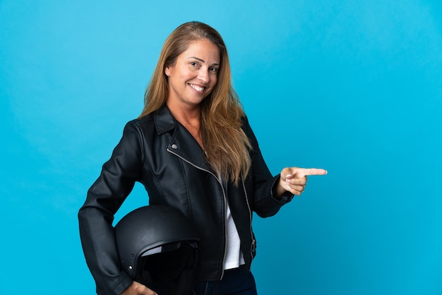 Middelbare leeftijd vrouw met een motorhelm geïsoleerd op blauwe muur wijzende vinger naar de zijkant