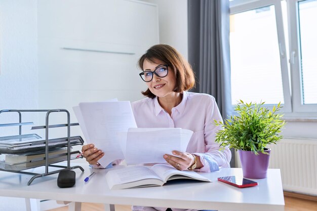 Middelbare leeftijd vrouw thuis werken aan een tafel met papieren Glimlachende volwassen vrouw in glazen kijken naar camera met zakelijke papieren documenten thuis werkplek onderwijs papierwerk concept