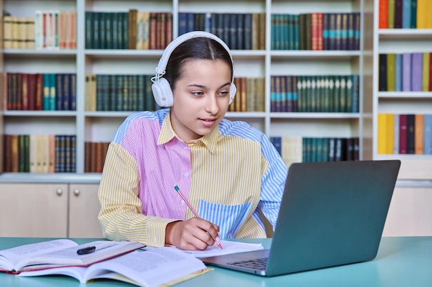 Middelbare school student meisje in koptelefoon studeren in schoolbibliotheek met behulp van boeken en laptop