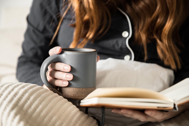 Middelsectie van een vrouw met een beker terwijl ze een boek op het bed leest