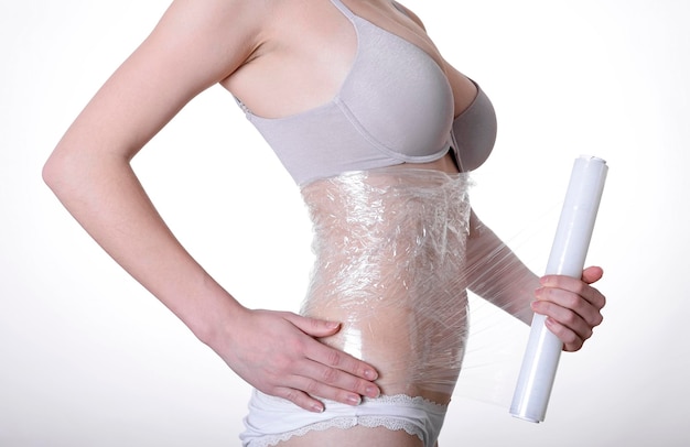 Middelsnede van een jonge vrouw die plastic om haar buik wikkelt tegen een witte achtergrond
