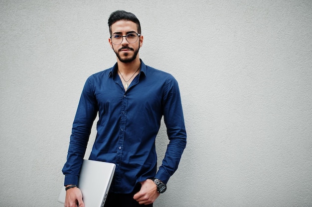 Midden-Oosterse ondernemer draagt een blauw shirt, een bril, houdt de laptop tegen een witte muur.