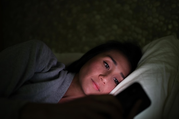 Middenvolwassen vrouw met een mobiele telefoon terwijl ze in de donkere kamer op bed ligt