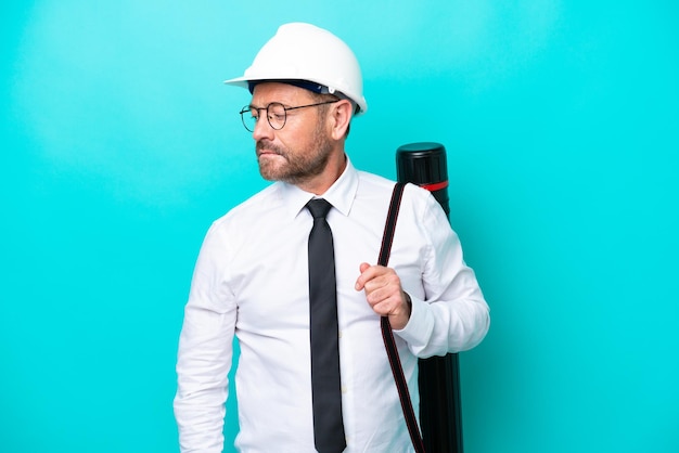 Middle age architect man with helmet and holding blueprints isolated on blue background looking to the side