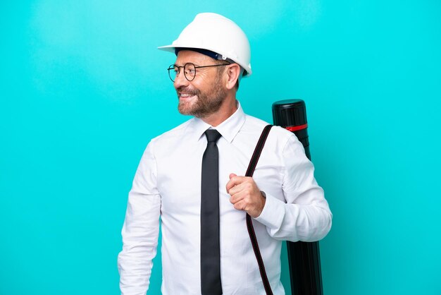 Middle age architect man with helmet and holding blueprints isolated on blue background looking side