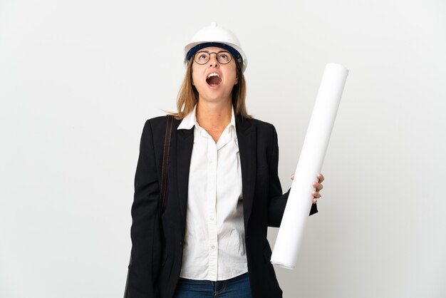 Middle age architect woman with helmet and holding blueprints over isolated looking up and with surprised expression