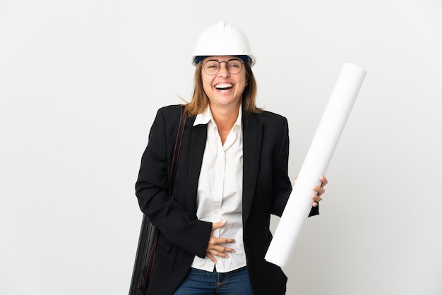 Middle age architect woman with helmet and holding blueprints over isolated wall smiling a lot