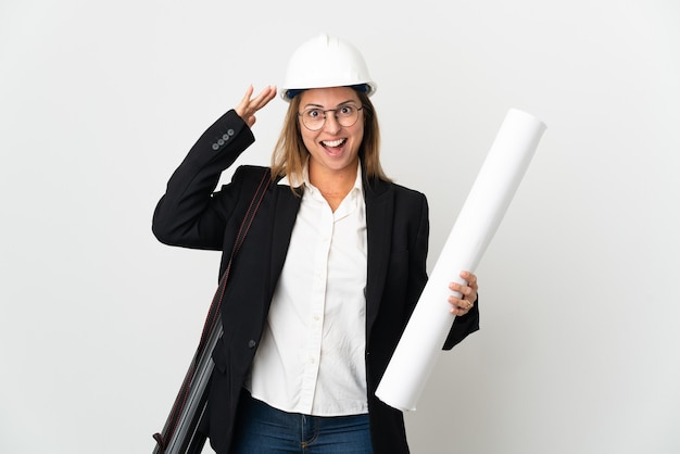 Middle age architect woman with helmet and holding blueprints over with surprise expression