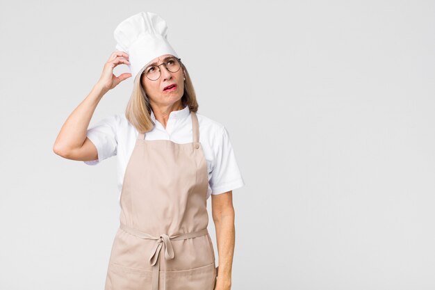 Foto donna del panettiere di mezza età sentirsi perplessa e confusa, grattandosi la testa e guardando di lato contro la parete piatta