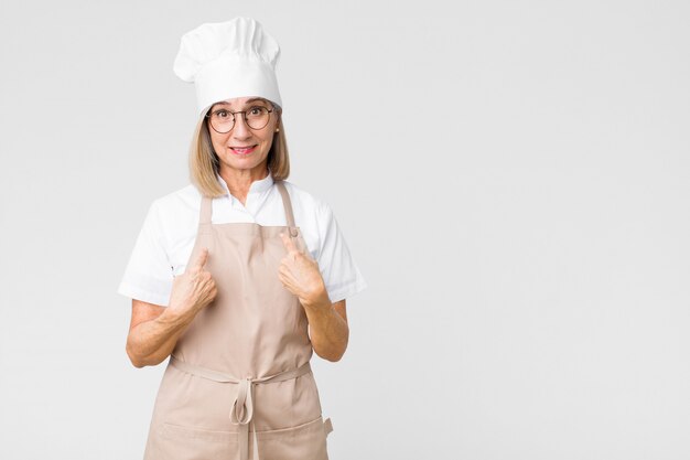 Middle age baker woman pointing to self with a confused and quizzical look, shocked and surprised to be chosen against flat wall