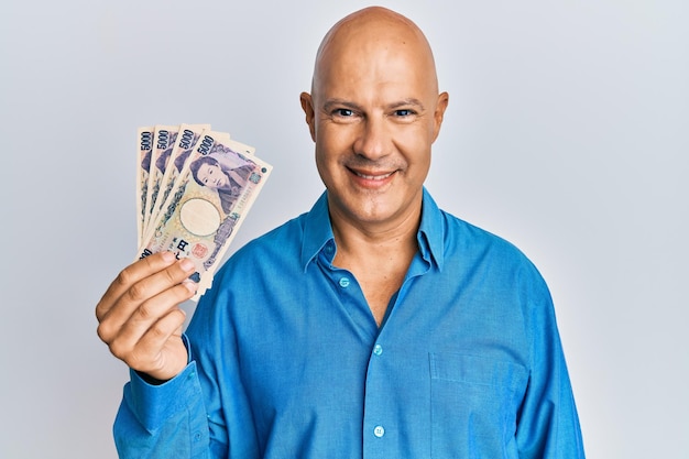 Лысый мужчина средних лет, держащий банкноты в 5000 японских иен, выглядит позитивно и счастливо, стоит и улыбается с уверенной улыбкой, показывая зубы