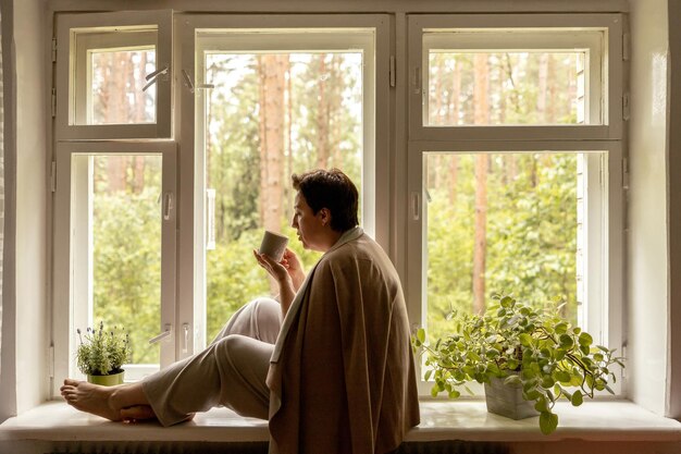 Middle age beautiful woman sitting on windowsill drinking tea dreaming 50yearold woman relaxing with cup of tea Relax at home looking away thinking stress free peaceful mood wellbeing alone