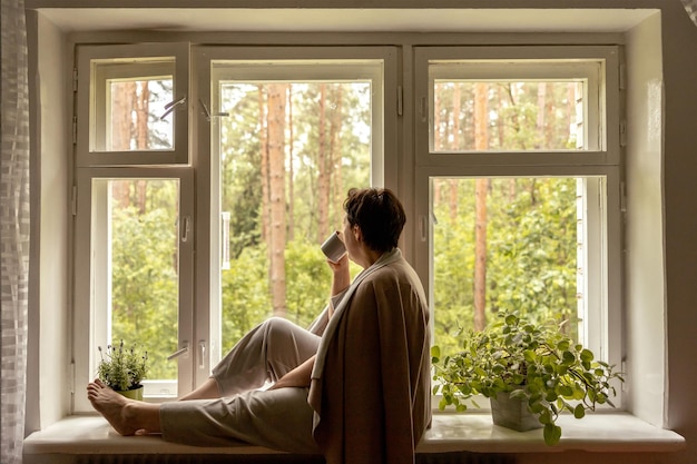 Middle age beautiful woman sitting on windowsill drinking tea dreaming 50yearold woman relaxing with cup of tea Relax at home looking away thinking stress free peaceful mood wellbeing alone